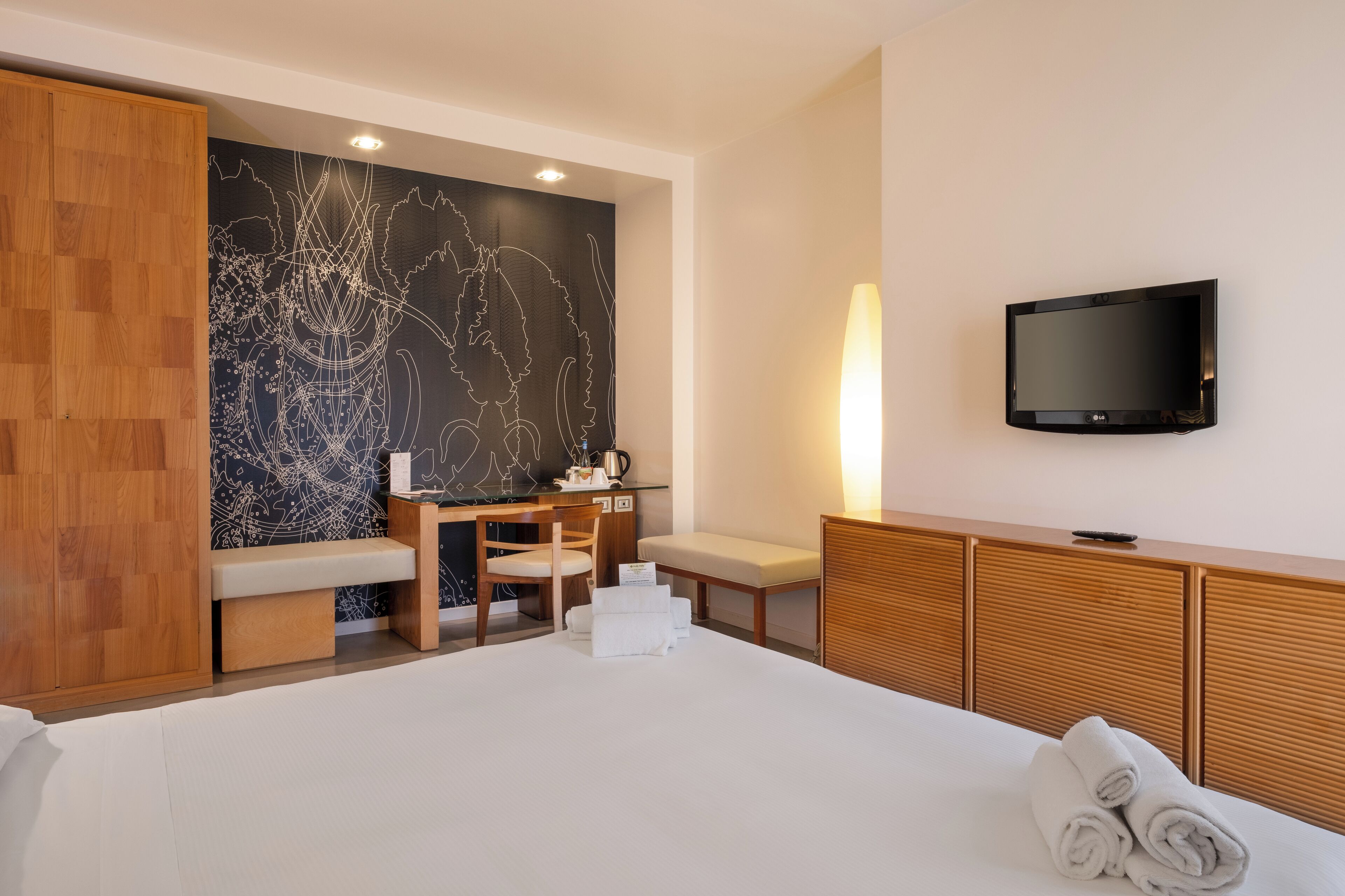 A hotel room with a large bed, a desk with a chair, a television mounted on the wall, and a wooden wardrobe.