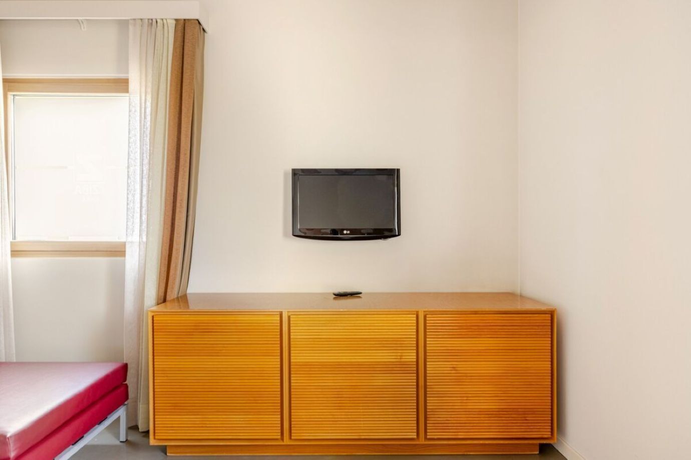 A room with a wooden cabinet, a television mounted on the wall, a window with curtains, and a red bench.