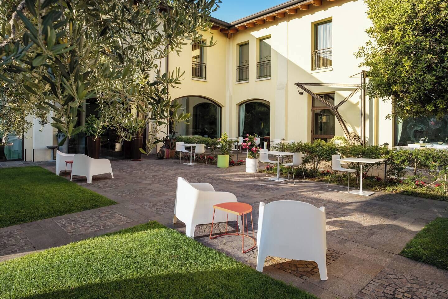 Un patio al aire libre con sillas y mesas blancas, rodeado de vegetación y un edificio con ventanales arqueados.