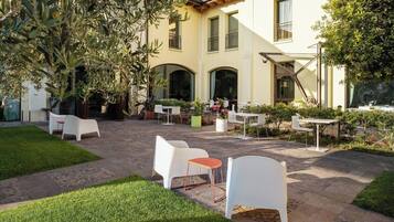 Un patio al aire libre con sillas y mesas blancas, rodeado de vegetación y un edificio con ventanales arqueados.