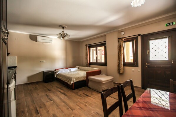 A room with a bed, a small table, and a chair. There is a window with curtains, a door, and a ceiling fan. - Alsos House (Kalabaka)