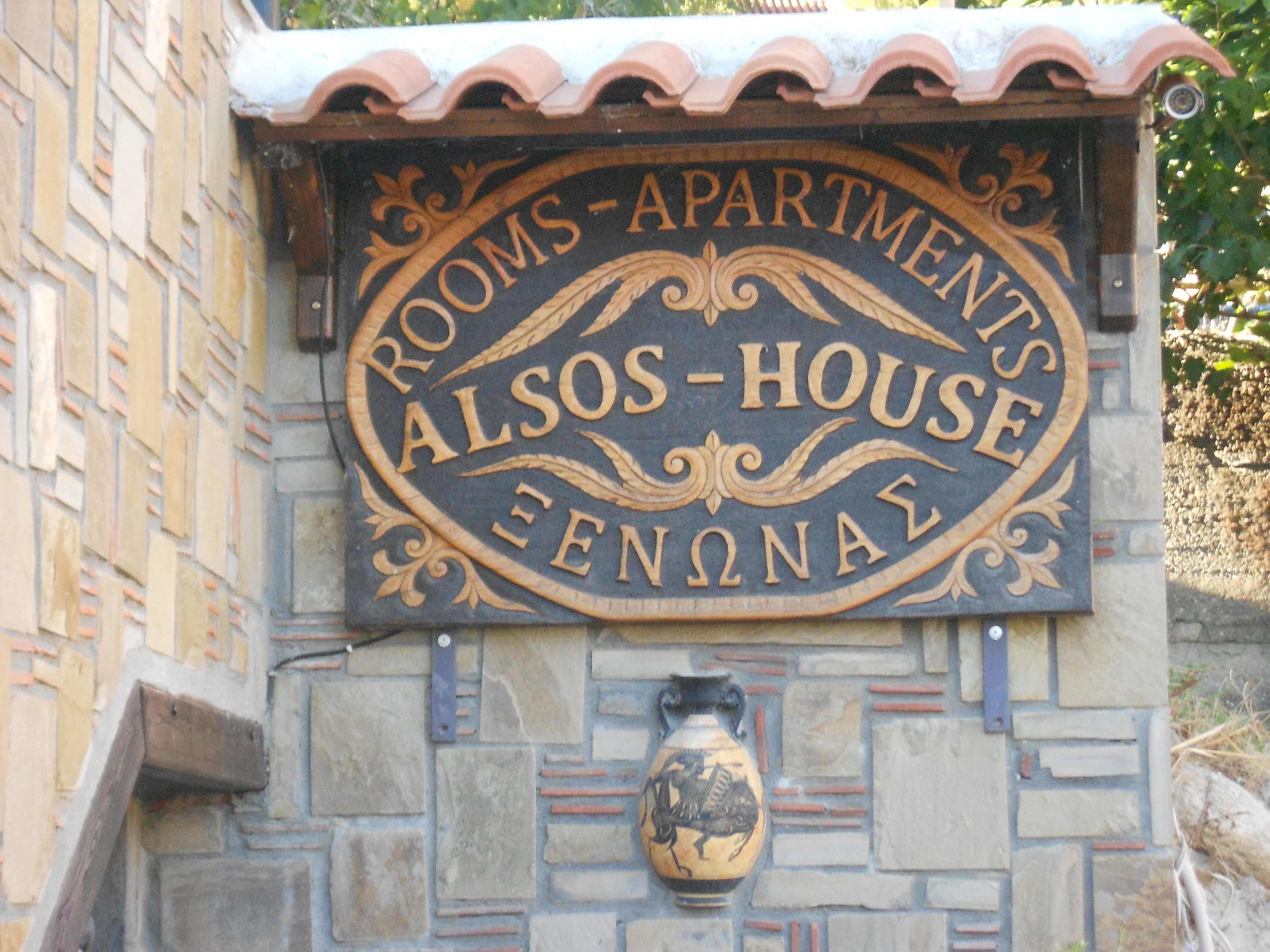 Une enseigne indiquant « Chambres – Appartements », avec le nom « Alos-House » et un vase décoratif en dessous.