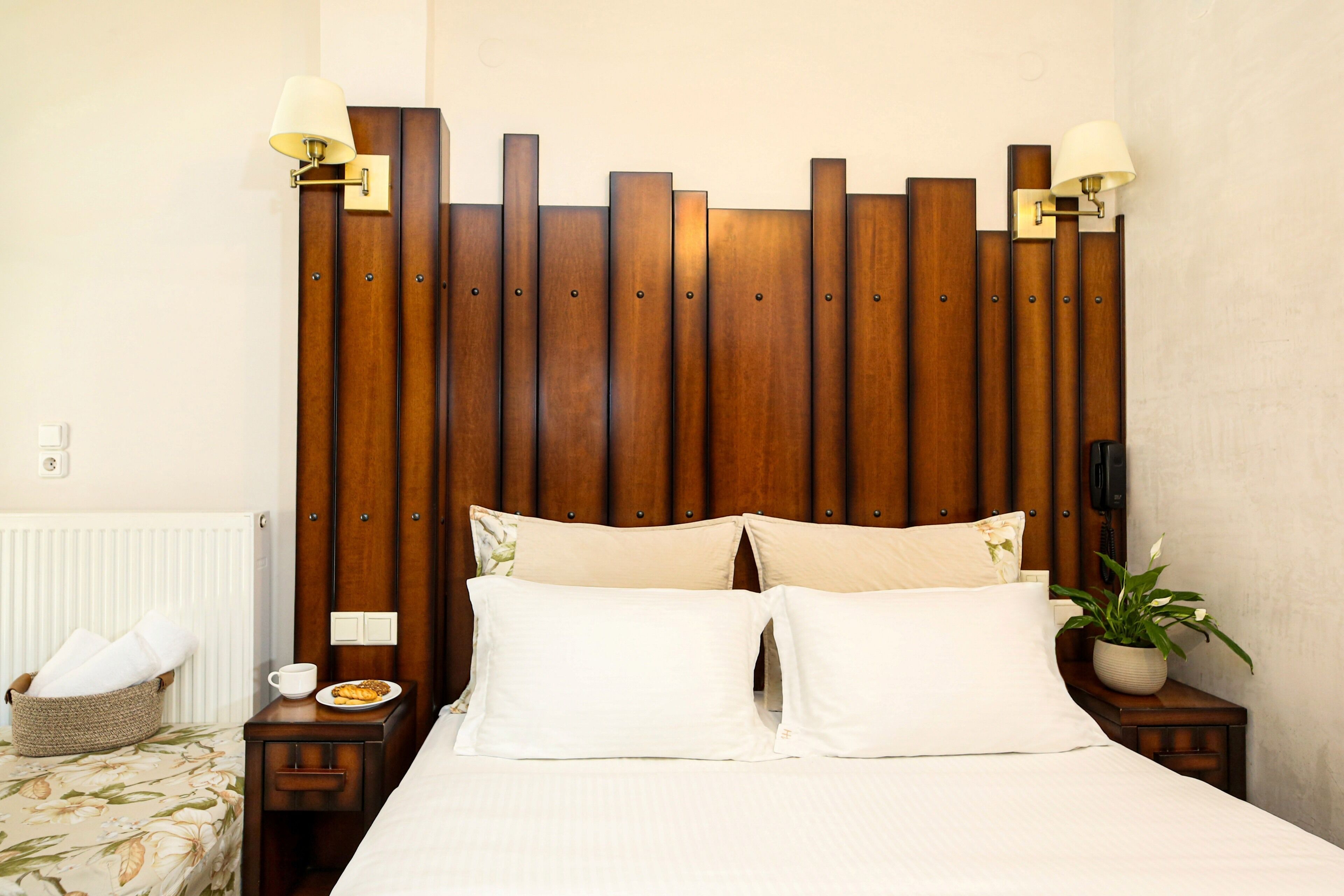 A hotel room with a wooden headboard, two bedside tables with lamps, a bed with white bedding, a small bedside table with a cup and plate, a basket with towels, and a potted plant.