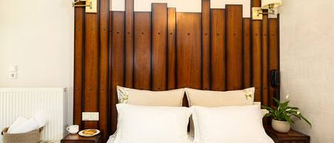 A hotel room with a wooden headboard, two bedside tables with lamps, a bed with white bedding, a small bedside table with a cup and plate, a basket with towels, and a potted plant.