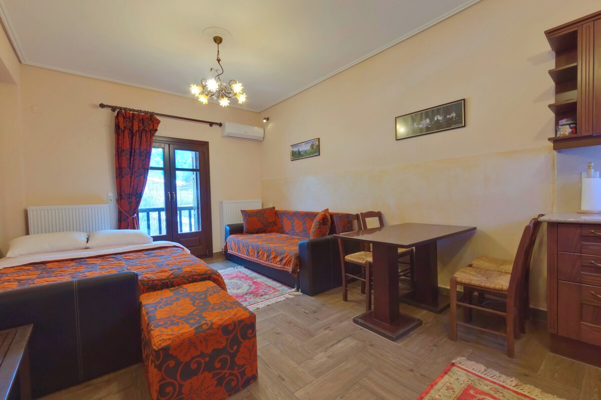 A room with a bed, a sofa, a dining table, and a chair. There is a window with curtains, a radiator, and a chandelier.