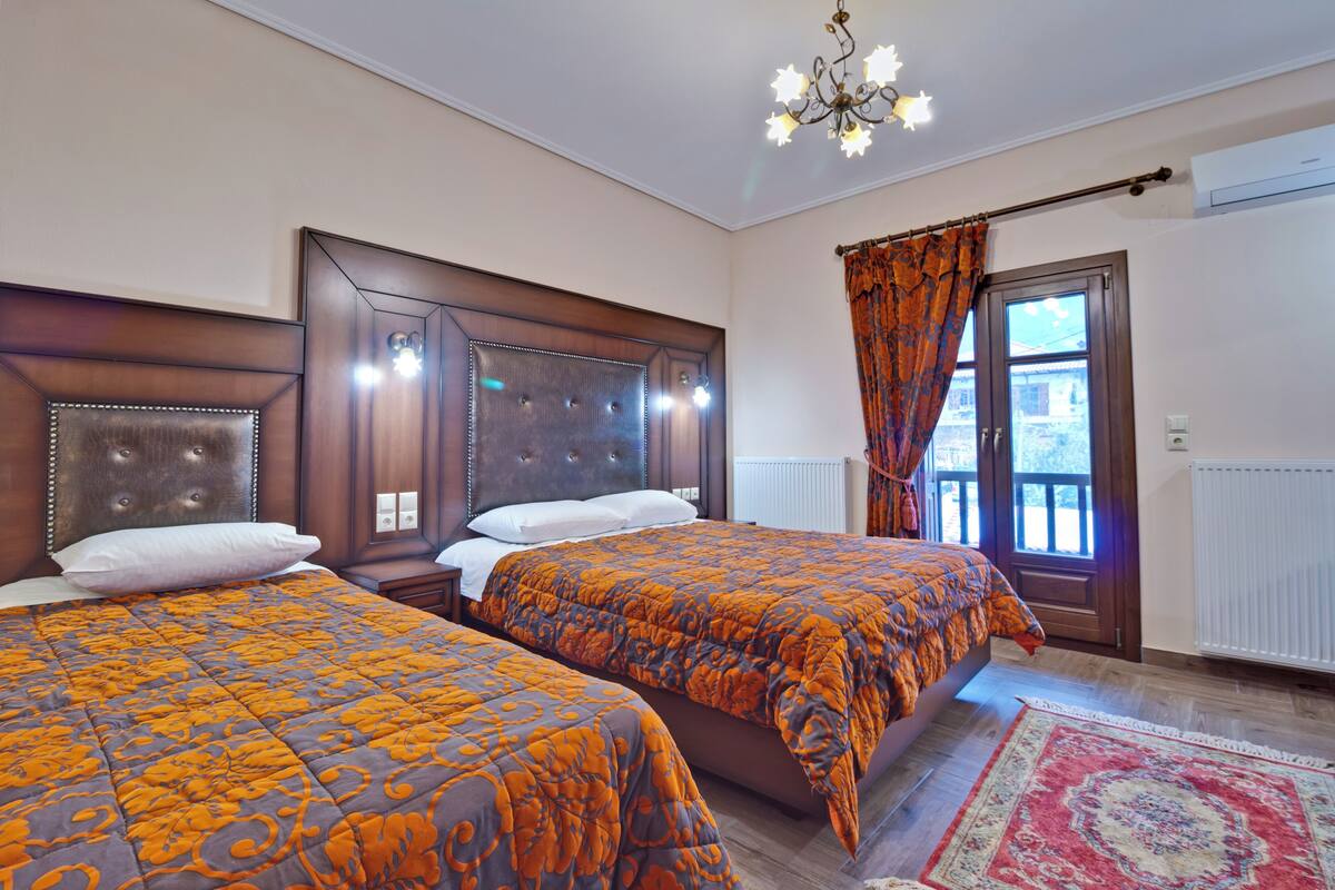 A hotel room with two beds, a chandelier, a window with curtains, and a radiator.