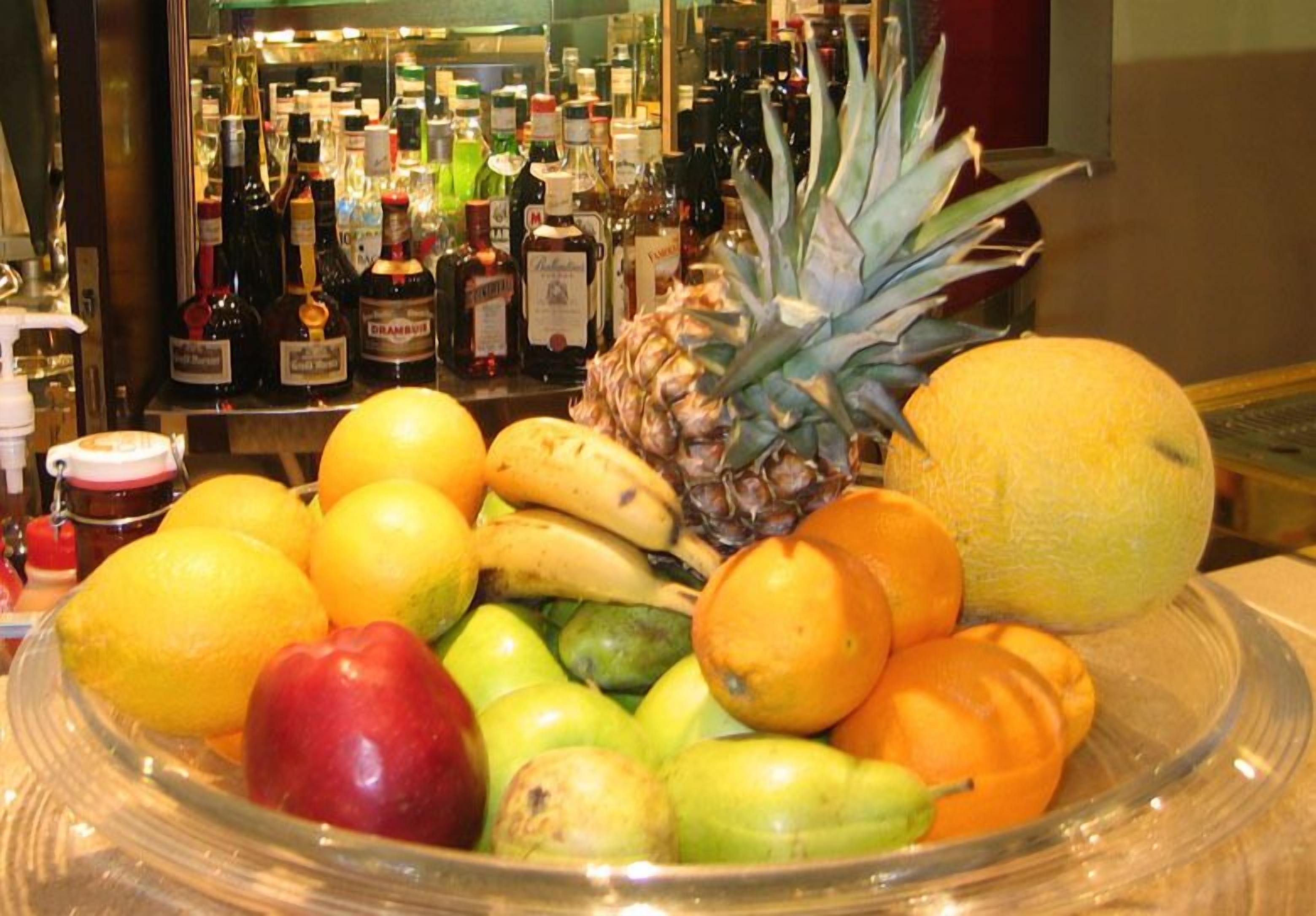 Uma tigela de frutas com diversas frutas e um bar com garrafas de bebidas ao fundo.