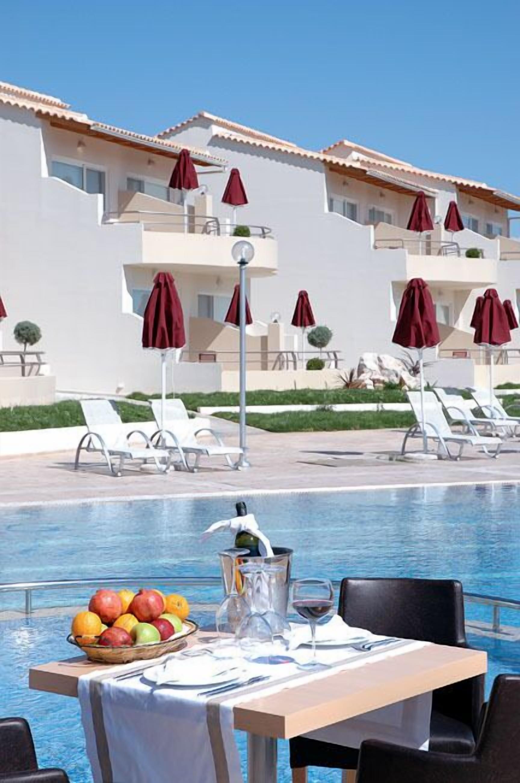 Zona de comedor junto a la piscina con una mesa preparada para dos personas, una cesta de fruta y una botella de vino. Se ven sillones de descanso y sombrillas al fondo.