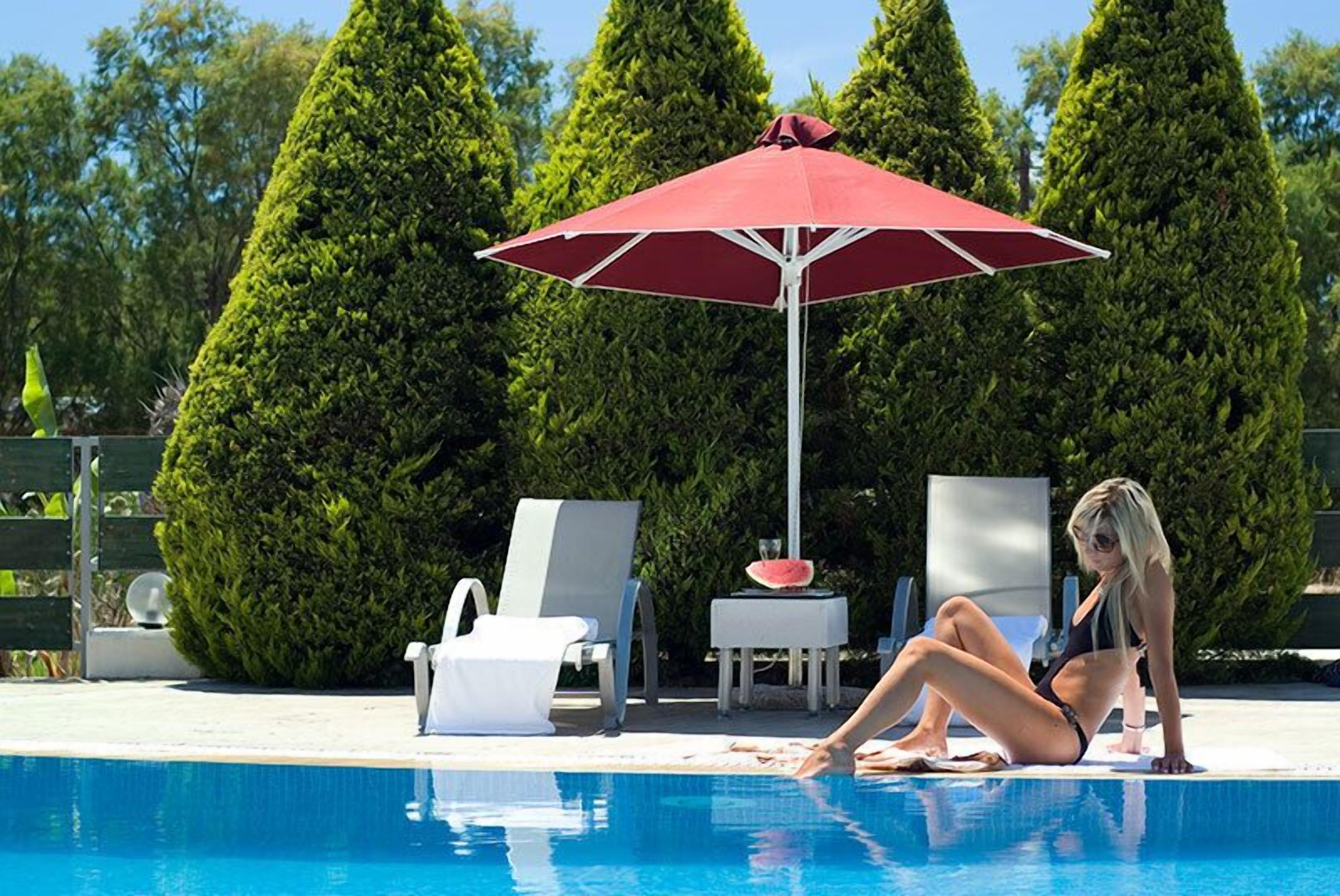 A poolside area with a red umbrella, lounge chairs, and a woman sitting by the pool.