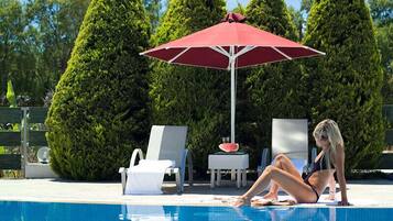 A poolside area with a red umbrella, lounge chairs, and a woman sitting by the pool.
