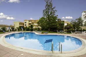 Piscina com espreguiçadeiras e uma grande árvore ao centro, cercada por edifícios.