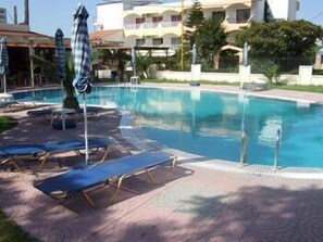 A swimming pool with lounge chairs and umbrellas, surrounded by a paved area and a building with balconies. - Lyristis Dinos Studios & Apartments (Rhodes)