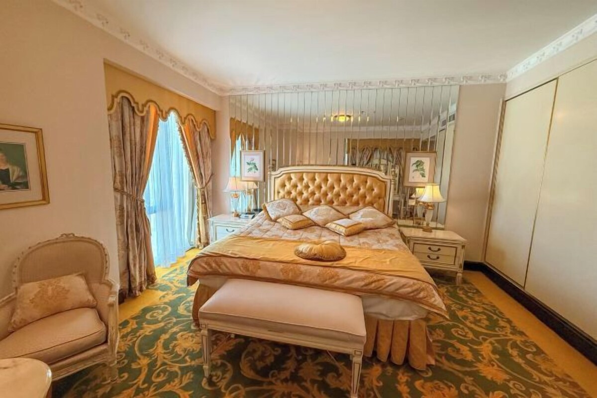A luxurious bedroom with a tufted headboard, a large bed with multiple pillows, a bench, a nightstand with a lamp, and a mirror.