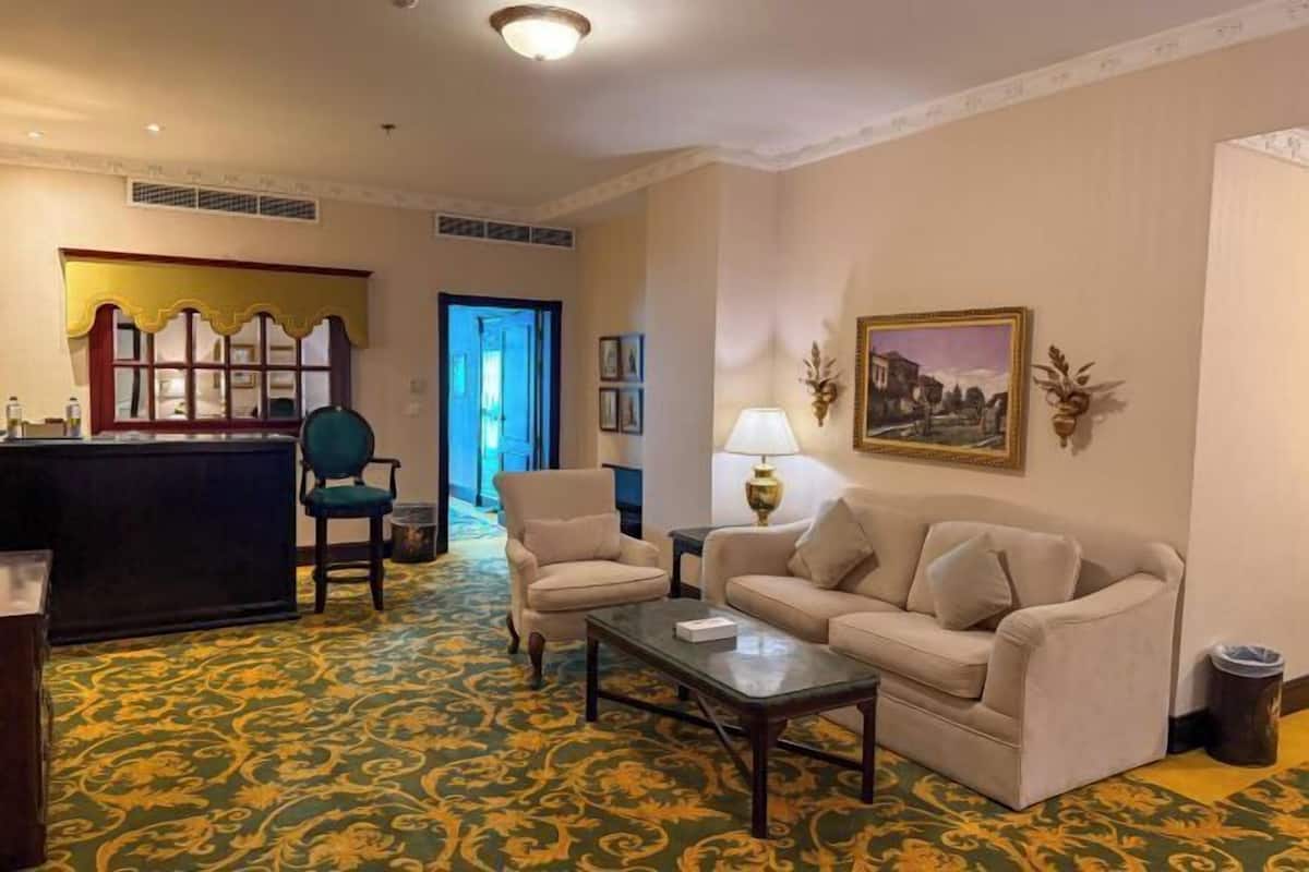 Een hotelkamer met een groen kamerbreed tapijt, een beige bank, twee fauteuils, een kleine tafel, een bureau met een stoel, een lamp en een ingelijst schilderij aan de muur.
