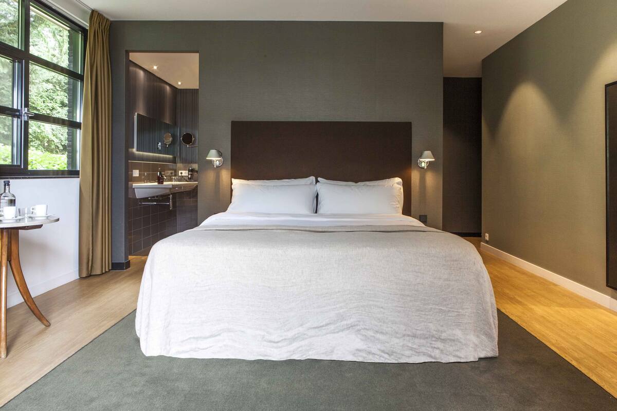 A modern hotel room with a large bed, a small table with a bottle and glasses, a bathroom with a mirror and sink, and a view of greenery through the window.