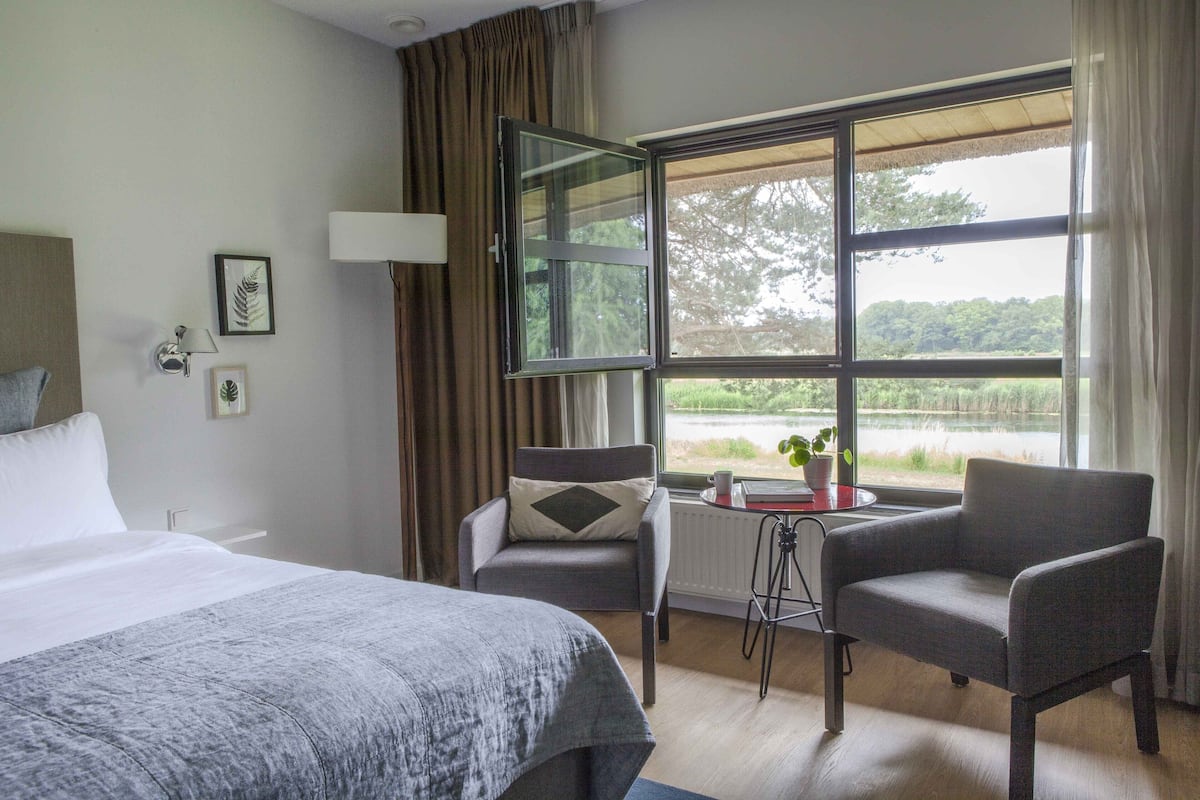 A hotel room with a bed, two armchairs, a small table with a plant, and a window with a view of greenery.