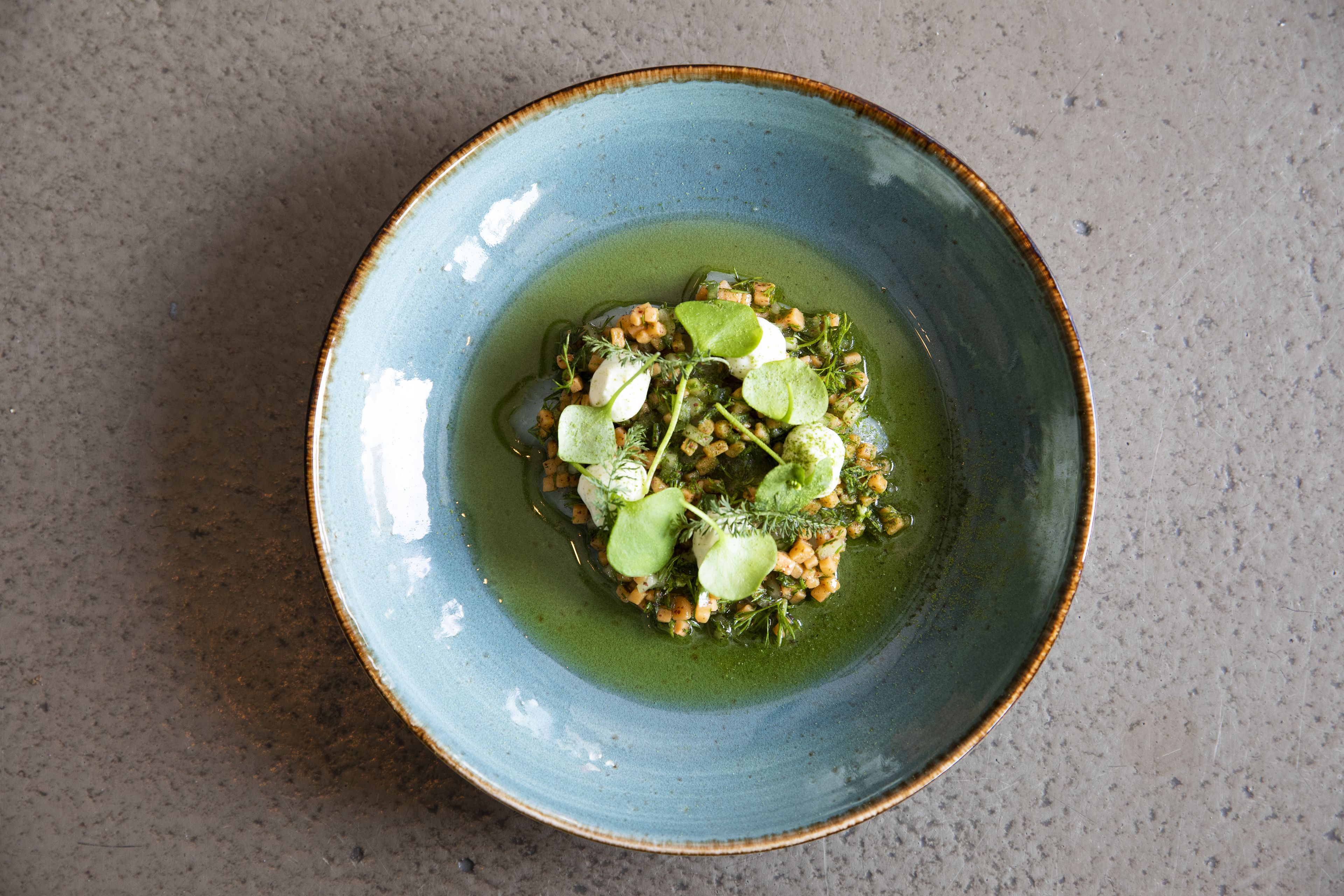 A blue ceramic bowl with a green sauce and garnishes on a textured surface.