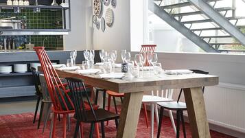 Een eethoek met een houten tafel gedekt voor zes personen, omringd door rode en zwarte stoelen. Een rood kleed bedekt de vloer, en in de achtergrond is een keuken zichtbaar met een zwart aanrecht en planken.