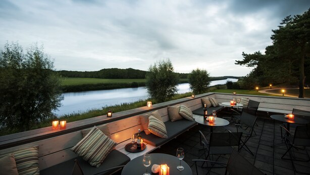An outdoor seating area with a view of a lake, furnished with sofas, tables, and candles. - Hotel Mooirivier (Dalfsen)