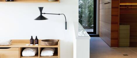Un baño moderno con estantes de madera, una lámpara de pie negra y vistas a árboles a través de un ventanal amplio.