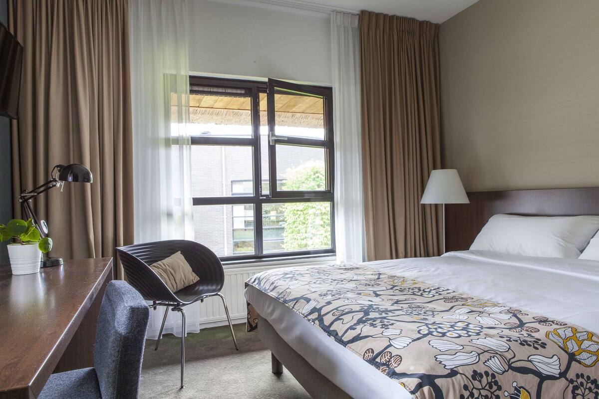 A hotel room with a bed, a desk with a chair, a window with curtains, and a lamp.