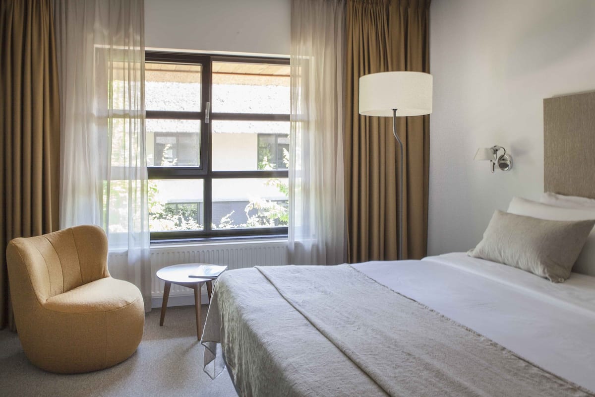 A hotel room with a bed, a chair, a small table, a lamp, and a window with curtains.