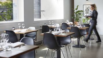 Un comedor moderno con mesas dispuestas para una comida, ventanas amplias y una planta en maceta.