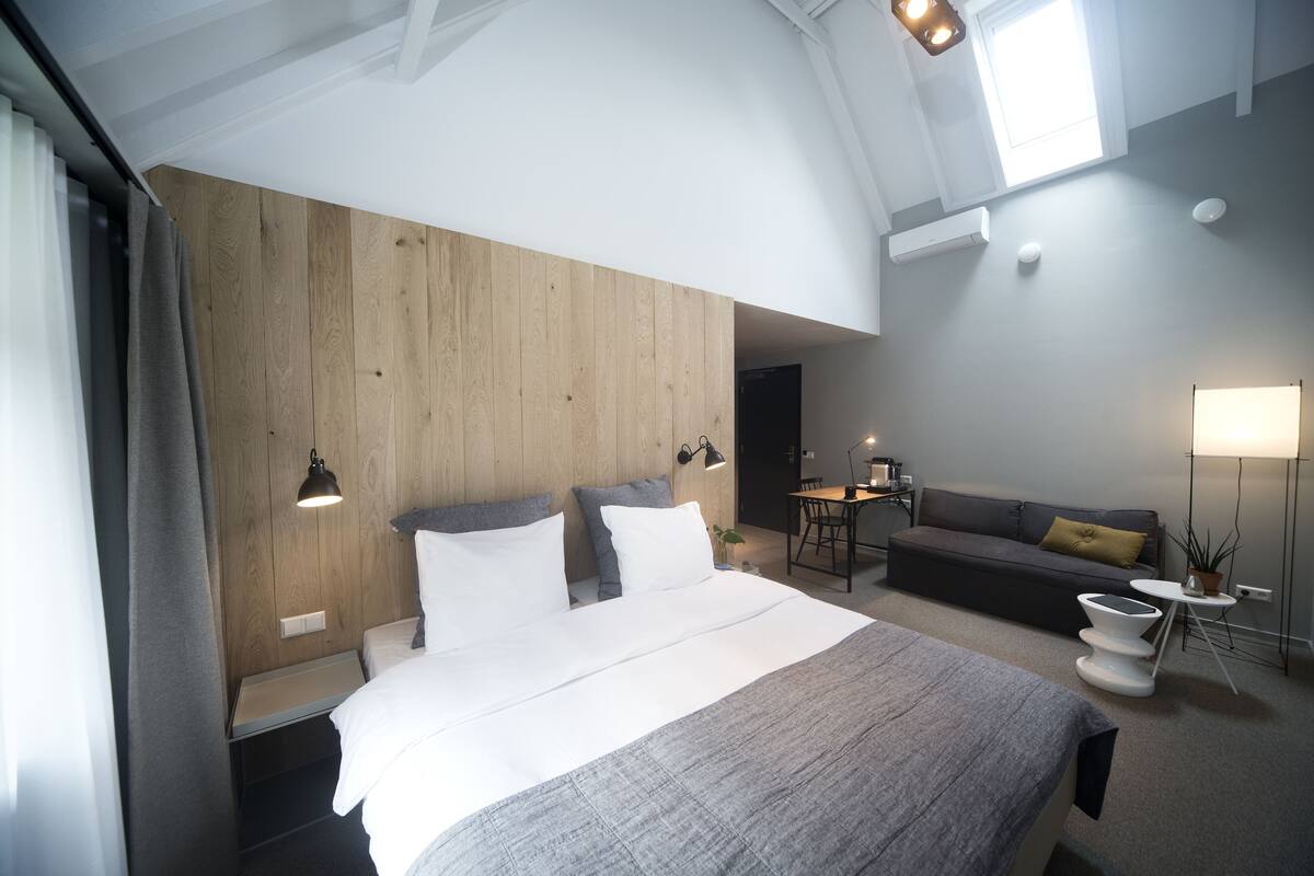 A modern hotel room with a large bed, wooden panel wall, and a small dining area.