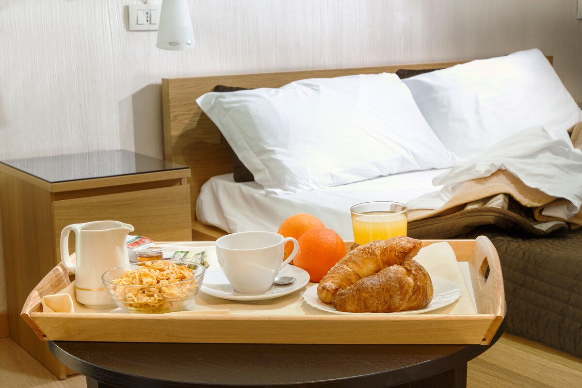 Una bandeja de desayuno ordenada con cruasanes, jugo de naranja, cereal y café sobre una mesa de madera en una habitación de hotel.