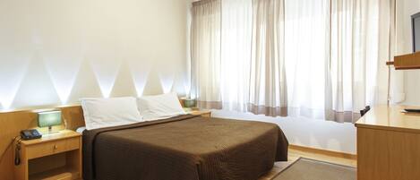 A hotel room with a bed, bedside table, lamp, and a window with curtains.
