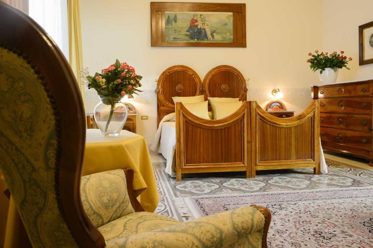 Un dormitorio tradicional con cama doble, cómoda de madera, mesita de noche con un jarrón de flores y alfombra estampada.