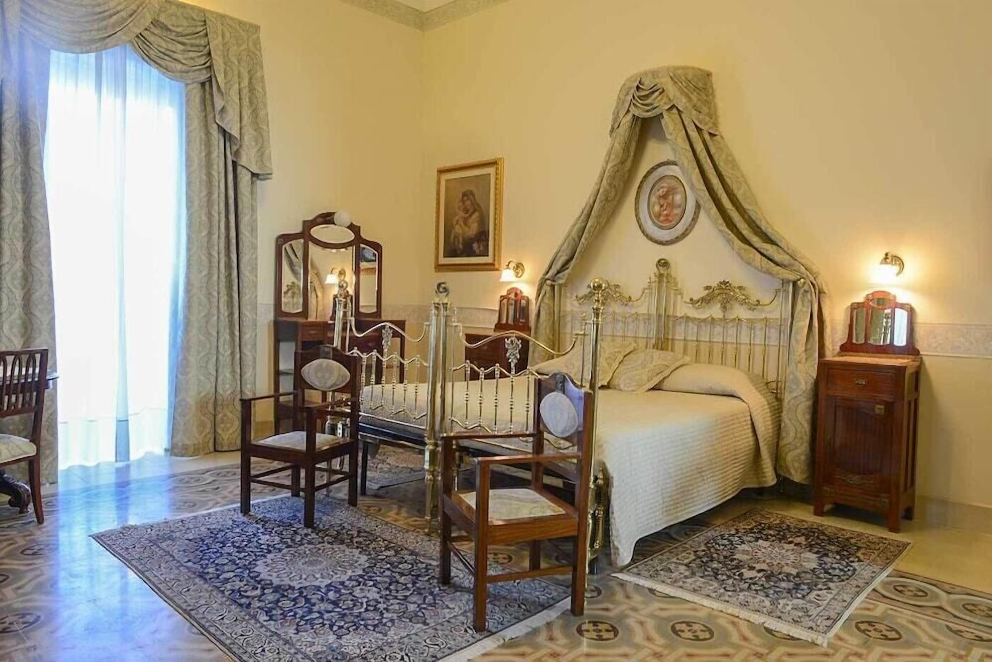 Quarto com cama de dossel, duas cadeiras de madeira, uma mesa de cabeceira e um espelho.
