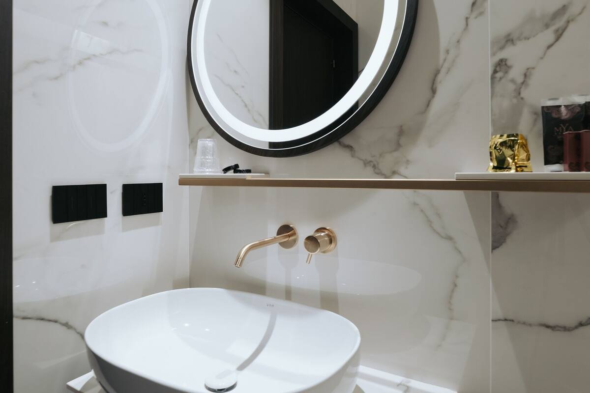 Un baño moderno con lavamanos de mármol blanco, grifería dorada y un espejo redondo con marco iluminado.