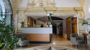 A reception area with a marble floor, a reception desk, two grey armchairs, and a large mural of a colonial building on the wall. - Albergo Italia (Matera)