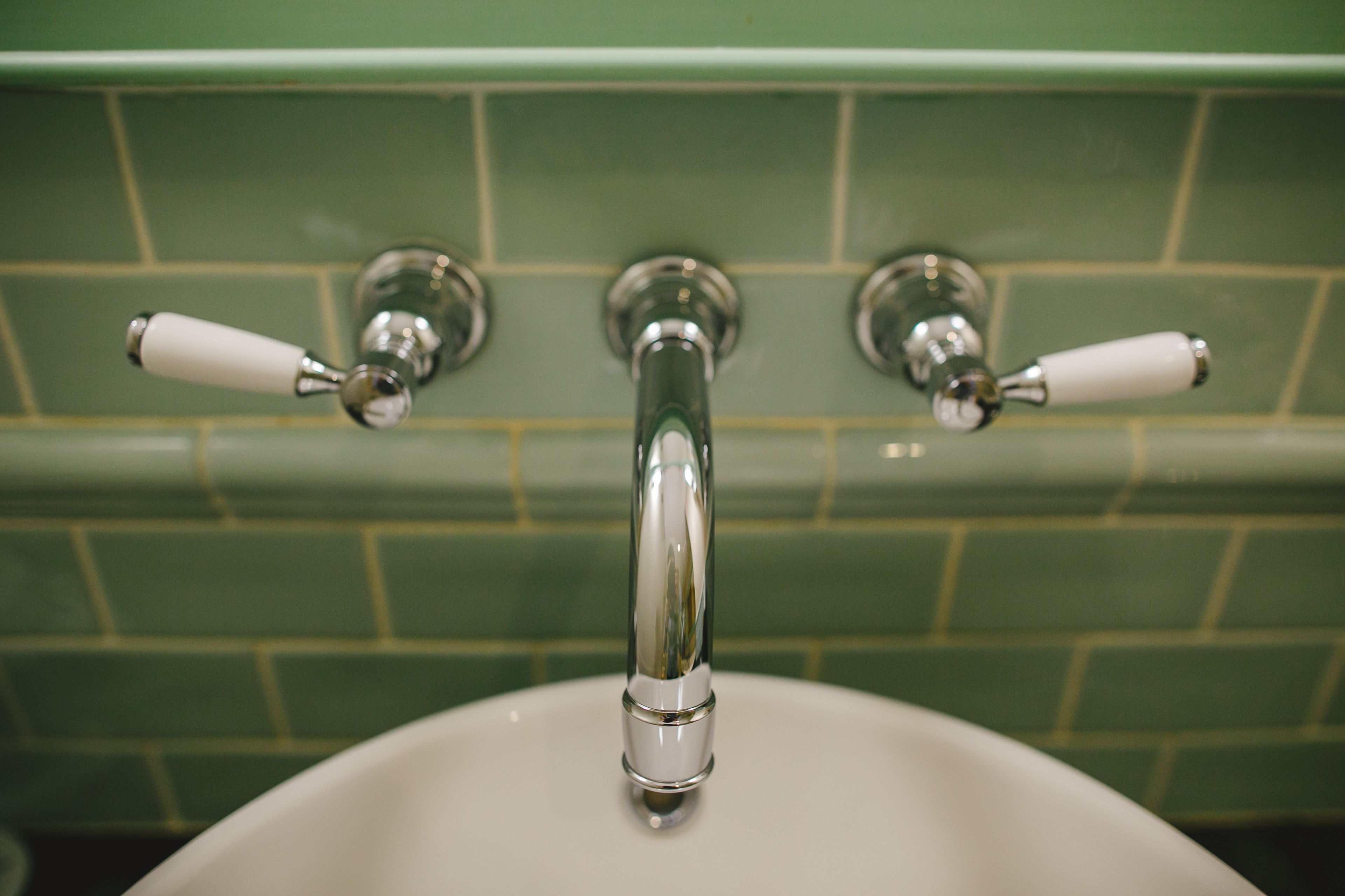 A bathroom with a white sink, green tiled walls, and three chrome faucets with white handles.