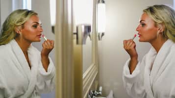 A person in a white bathrobe applying lipstick in front of a mirror.