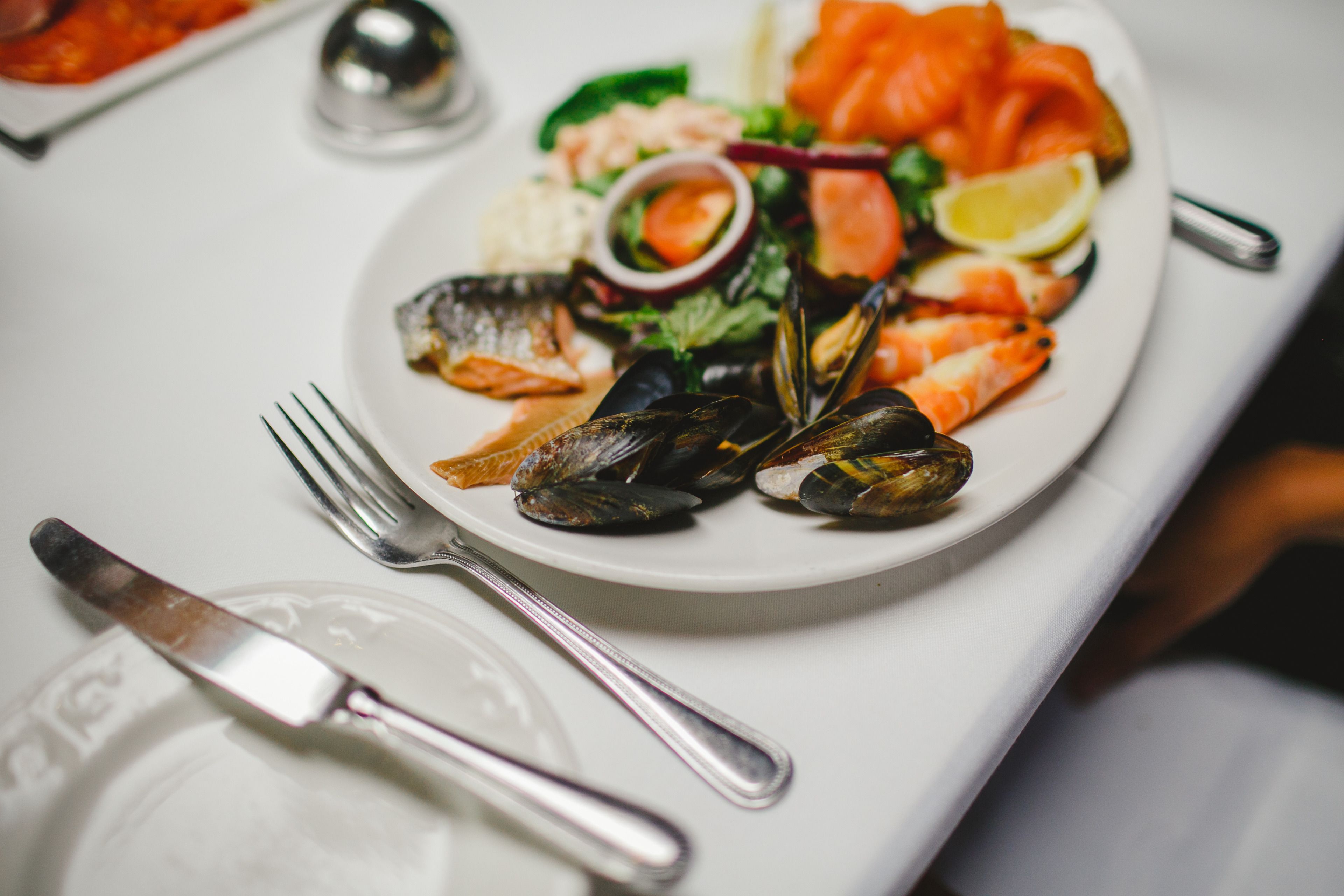 A seafood platter with mussels, shrimp, and salmon on a white plate, accompanied by a fork and knife on a table.