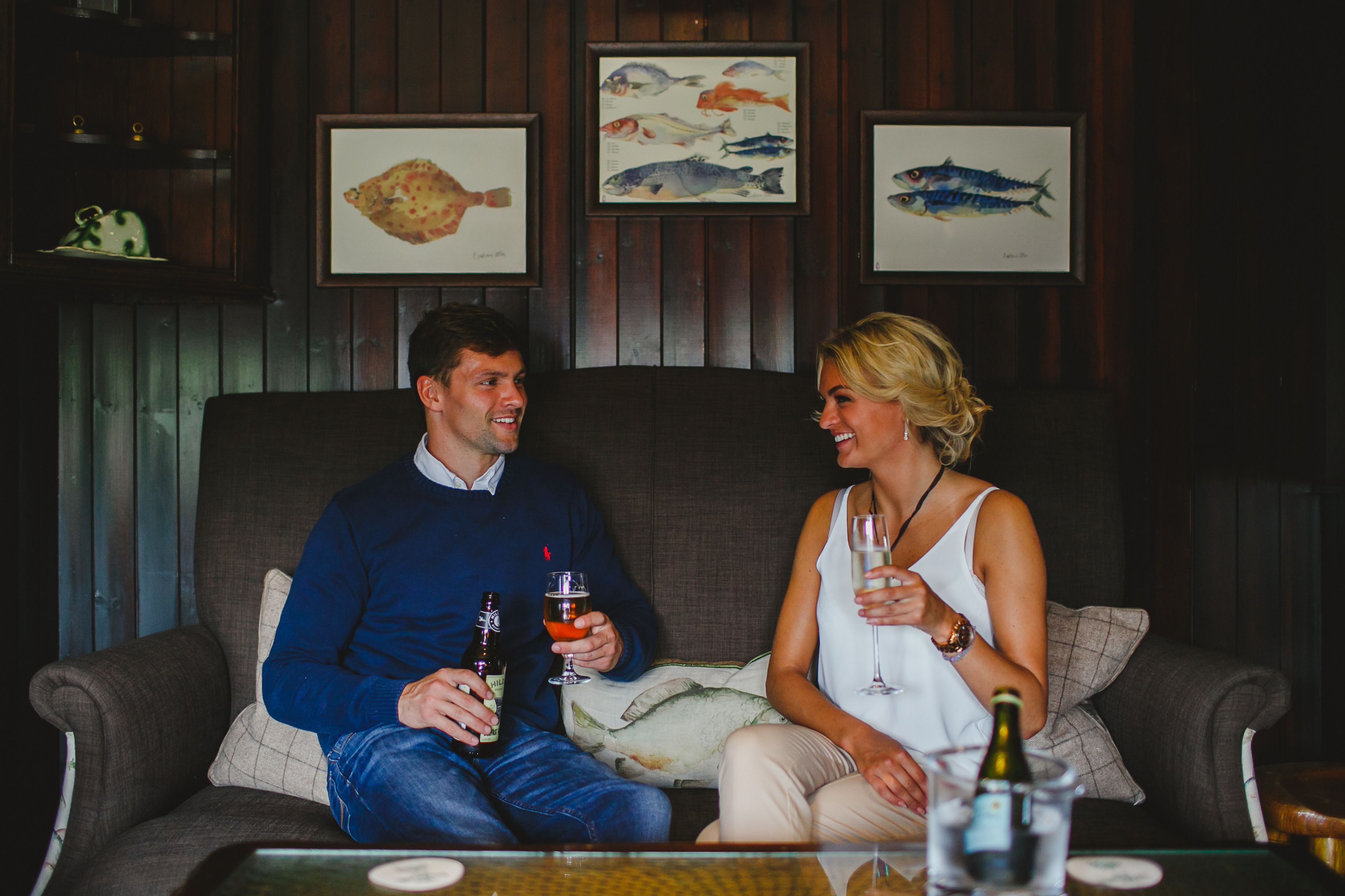A couple sitting on a couch in a room with wooden paneling and framed fish artwork on the wall.