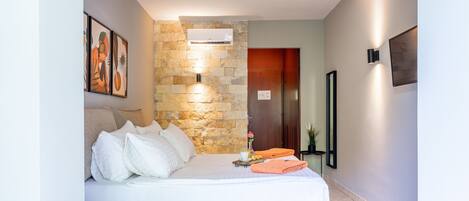 Una habitación de hotel moderna con una pared de acento de piedra, una cama con ropa de cama blanca, una mesita de noche y una pequeña mesa con un jarrón y una toalla naranja.