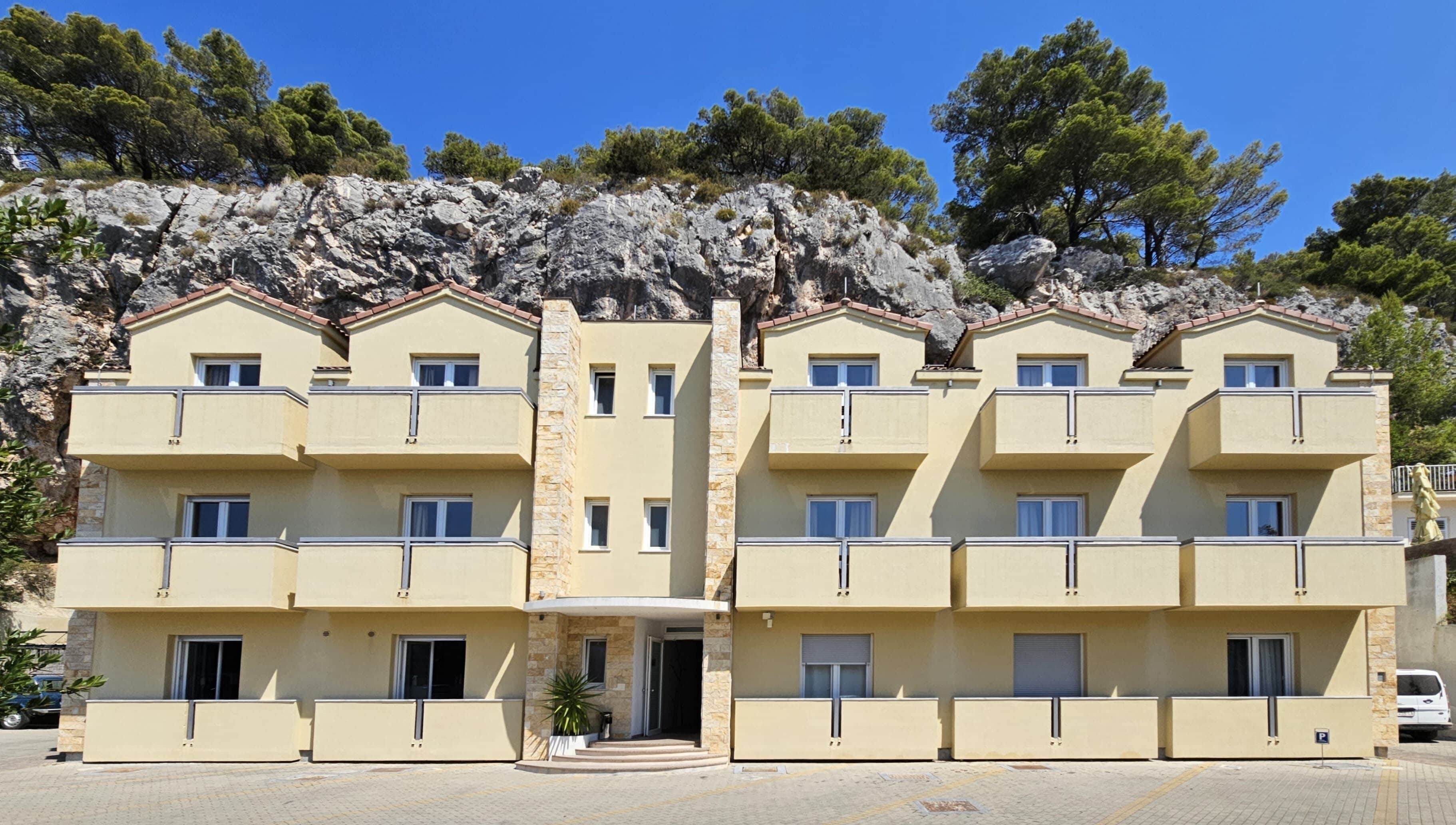 A multi-story building with balconies, a stone facade, and a parking area.