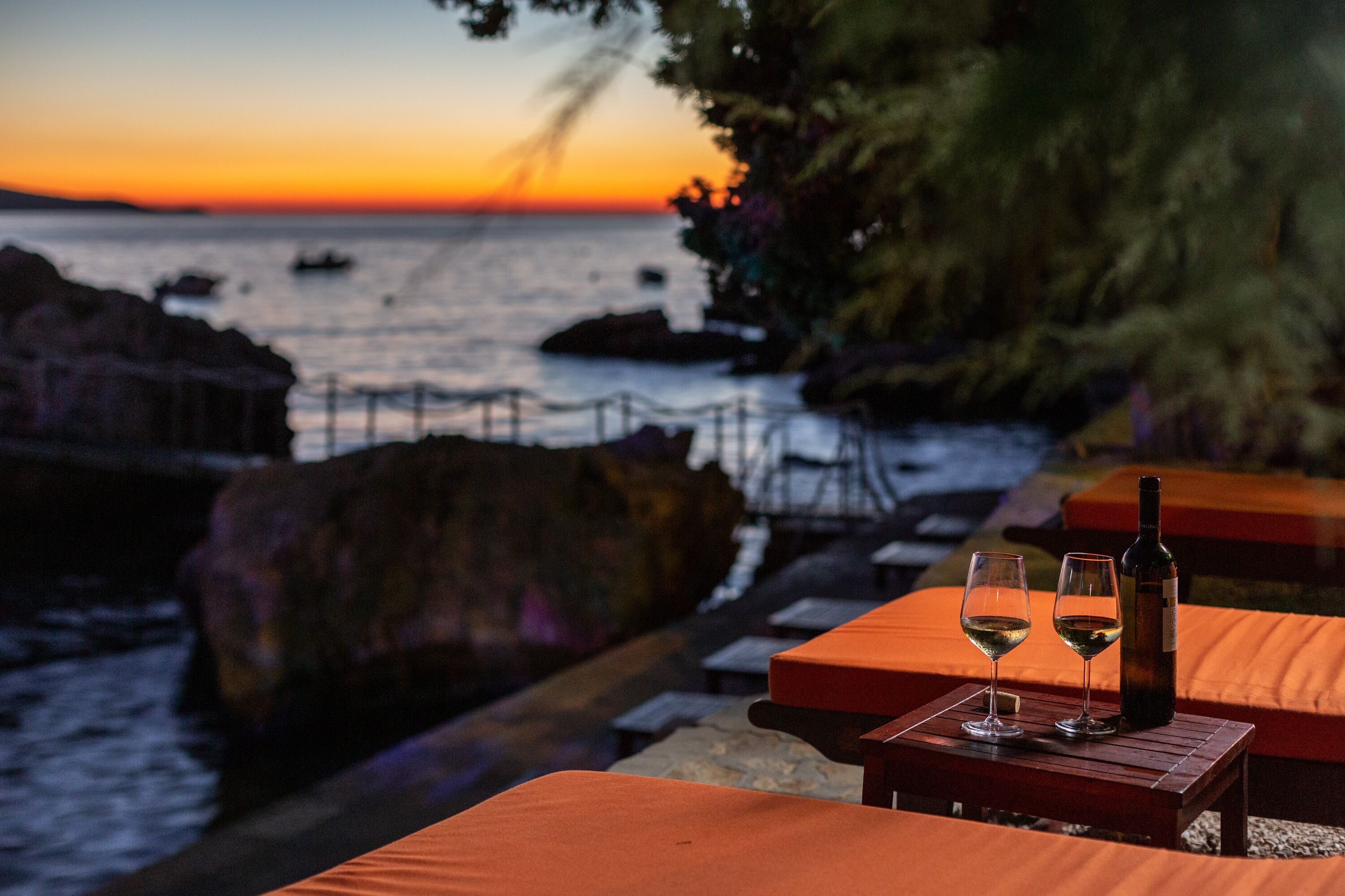 A sunset view from a seaside restaurant with two wine glasses and a bottle on a wooden table.