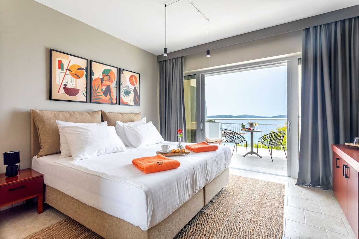 A modern hotel room with a large bed, a balcony with a table and chairs, and artwork on the wall.