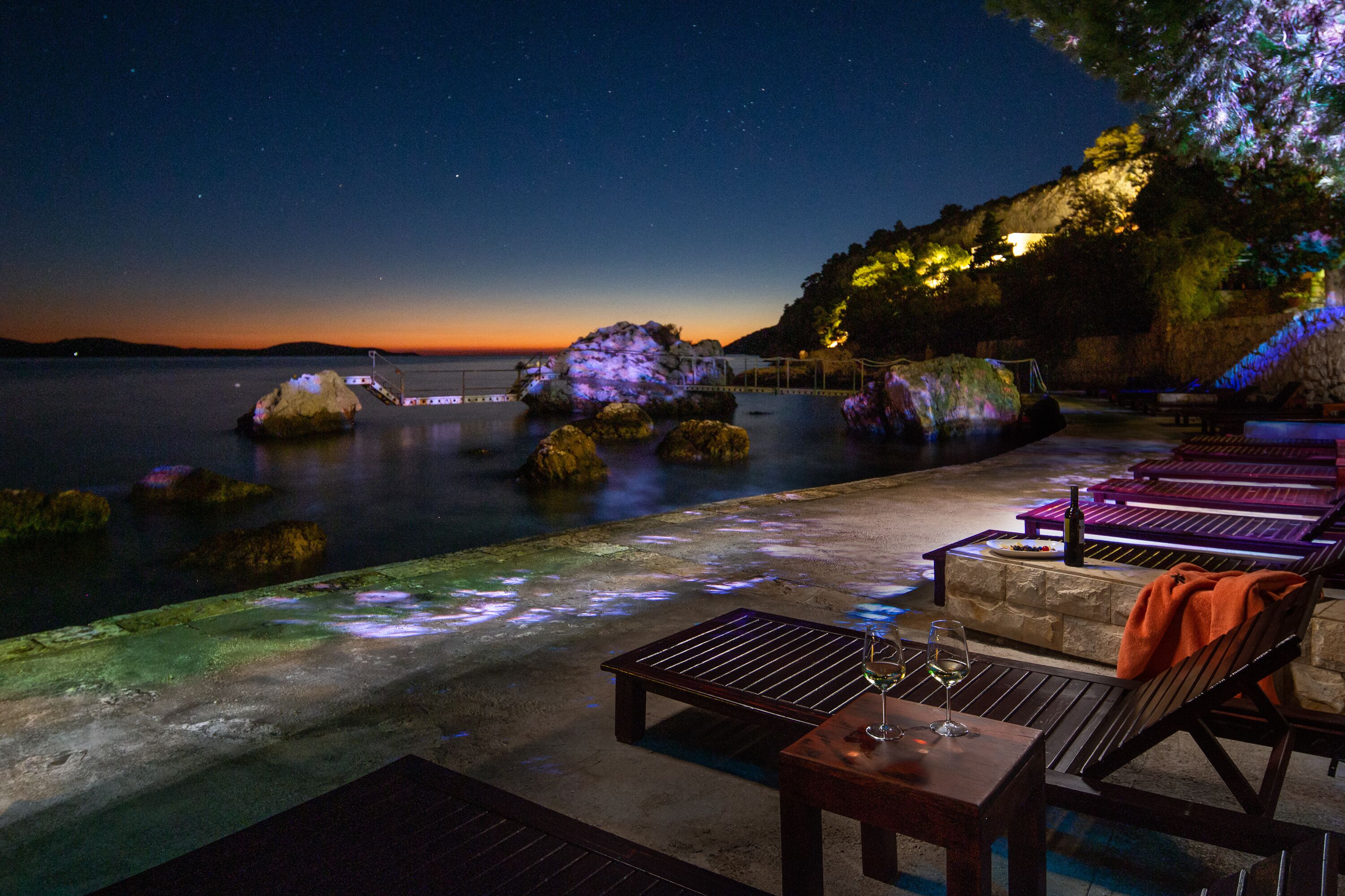 A waterfront patio with seating and tables, overlooking a calm body of water and a starry night sky.