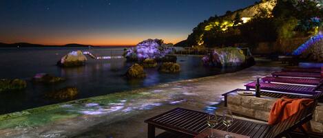 Un patio frente al agua con asientos y mesas, con vista a una superficie acuática tranquila y un cielo estrellado.