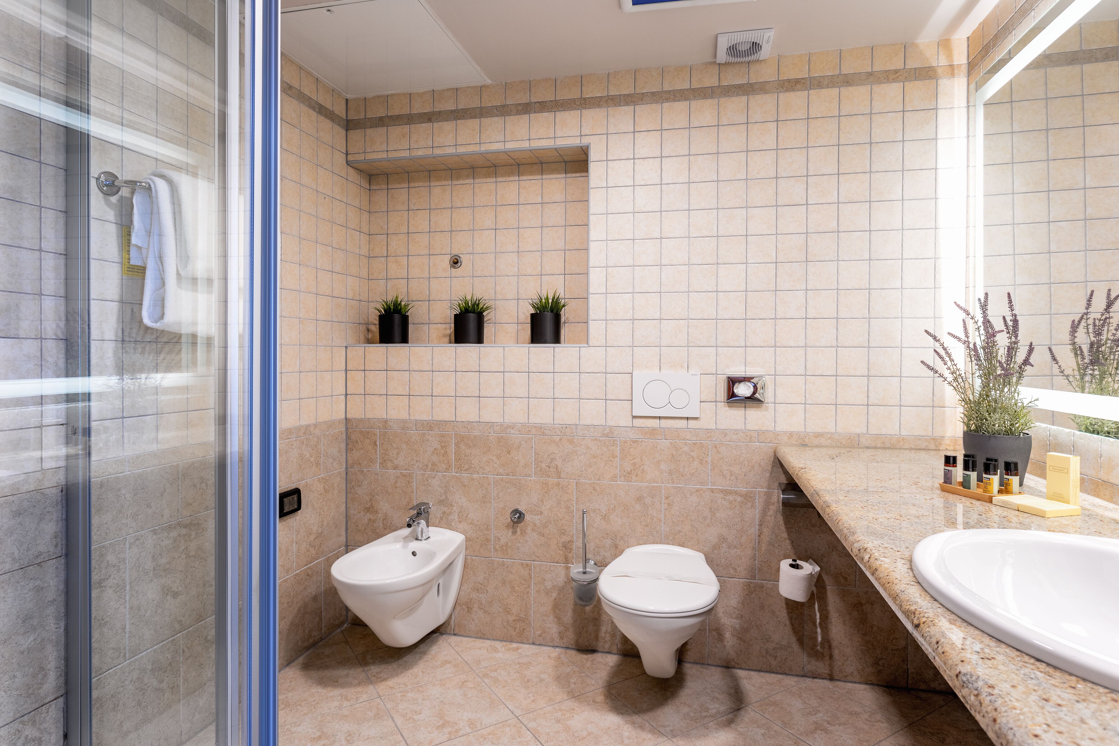 A bathroom with a glass shower enclosure, a bidet, a toilet, and a sink with a marble countertop.
