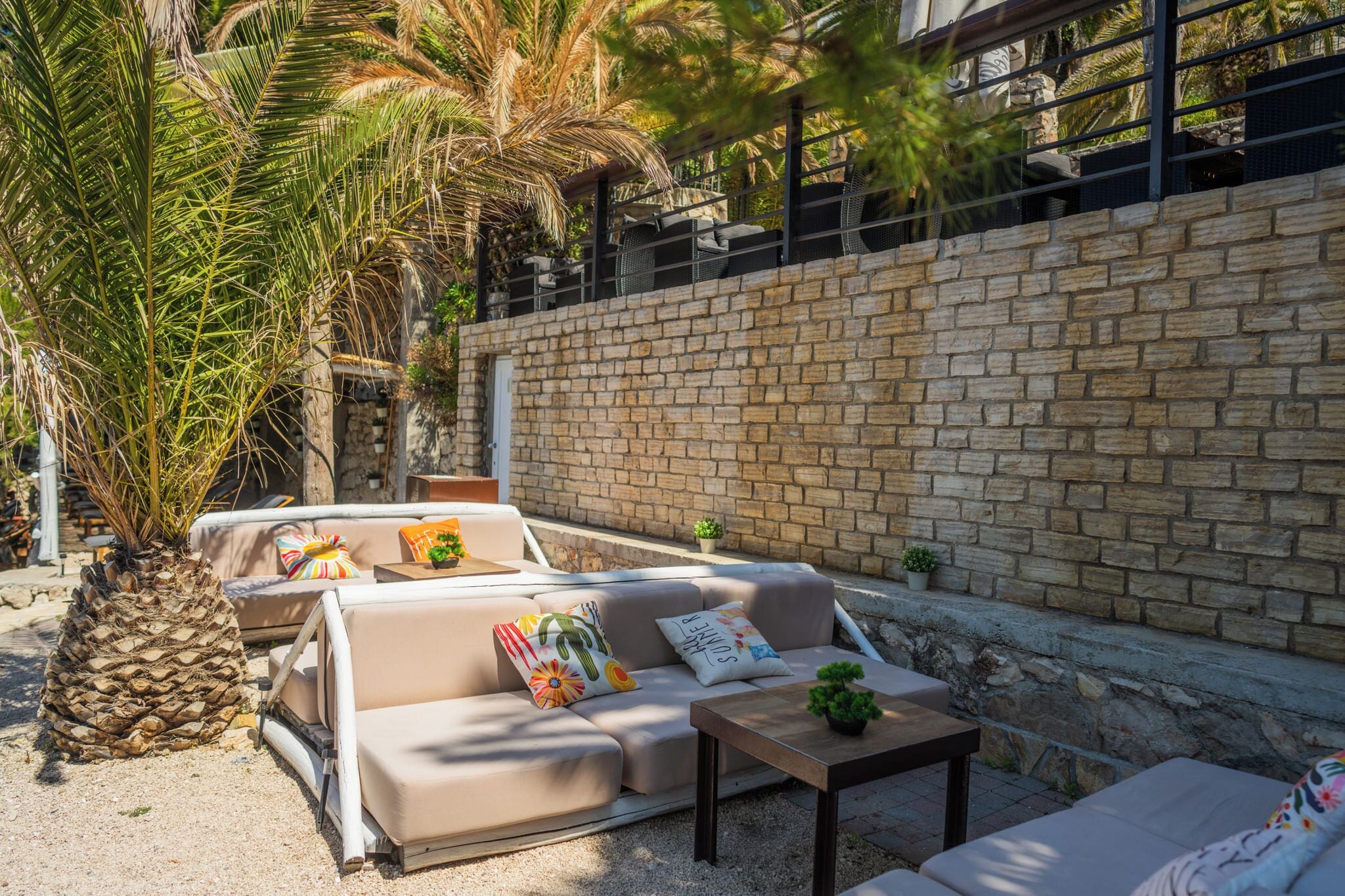 An outdoor seating area with a stone wall, a palm tree, and a cushioned sofa with decorative pillows.
