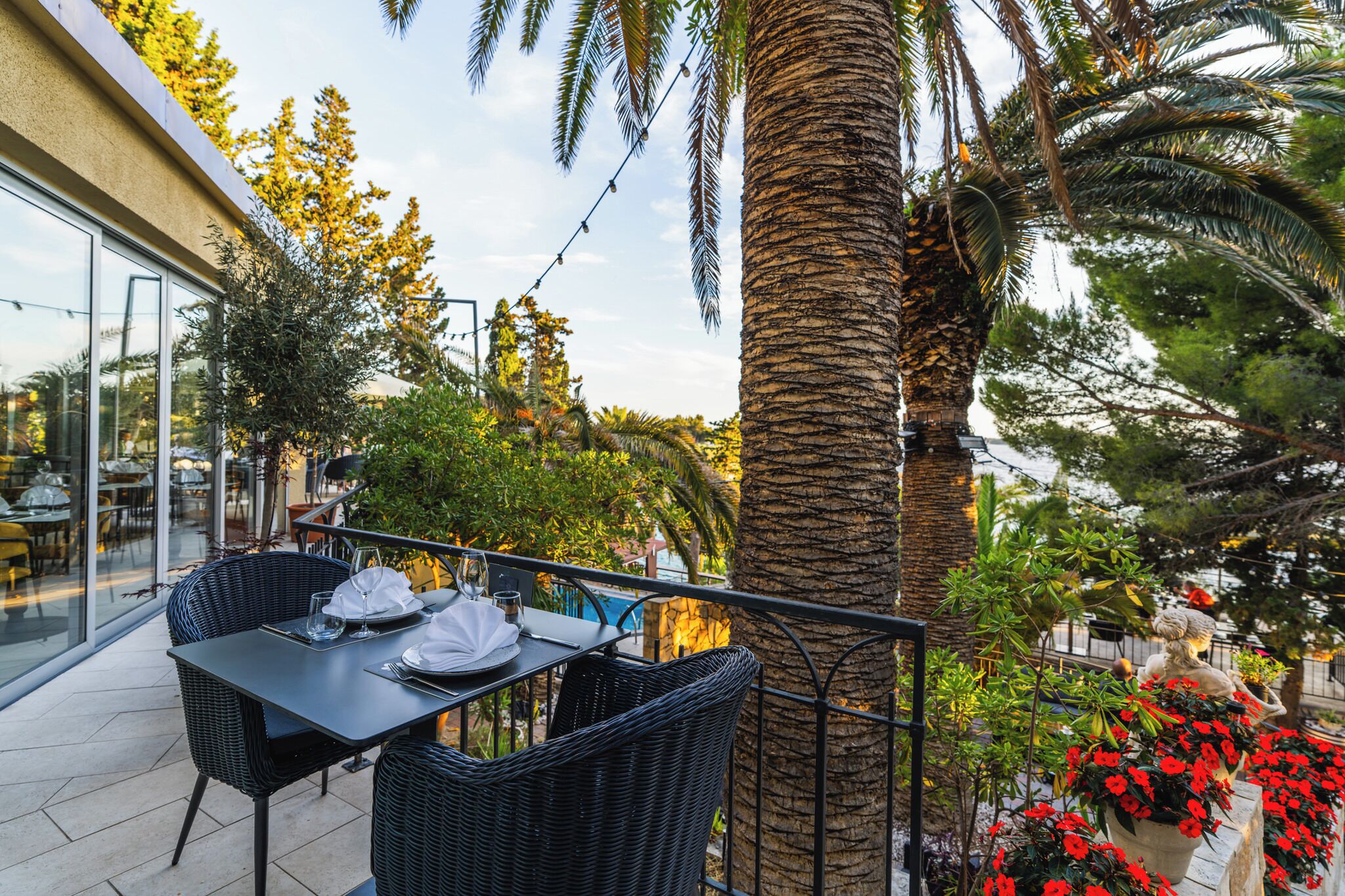 An outdoor dining area with wicker chairs and a table set for two, surrounded by palm trees and potted plants.