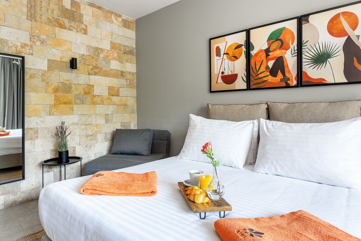 A hotel room with a stone wall, a bed with white linens, a gray sofa, and a breakfast tray with croissants and orange juice.