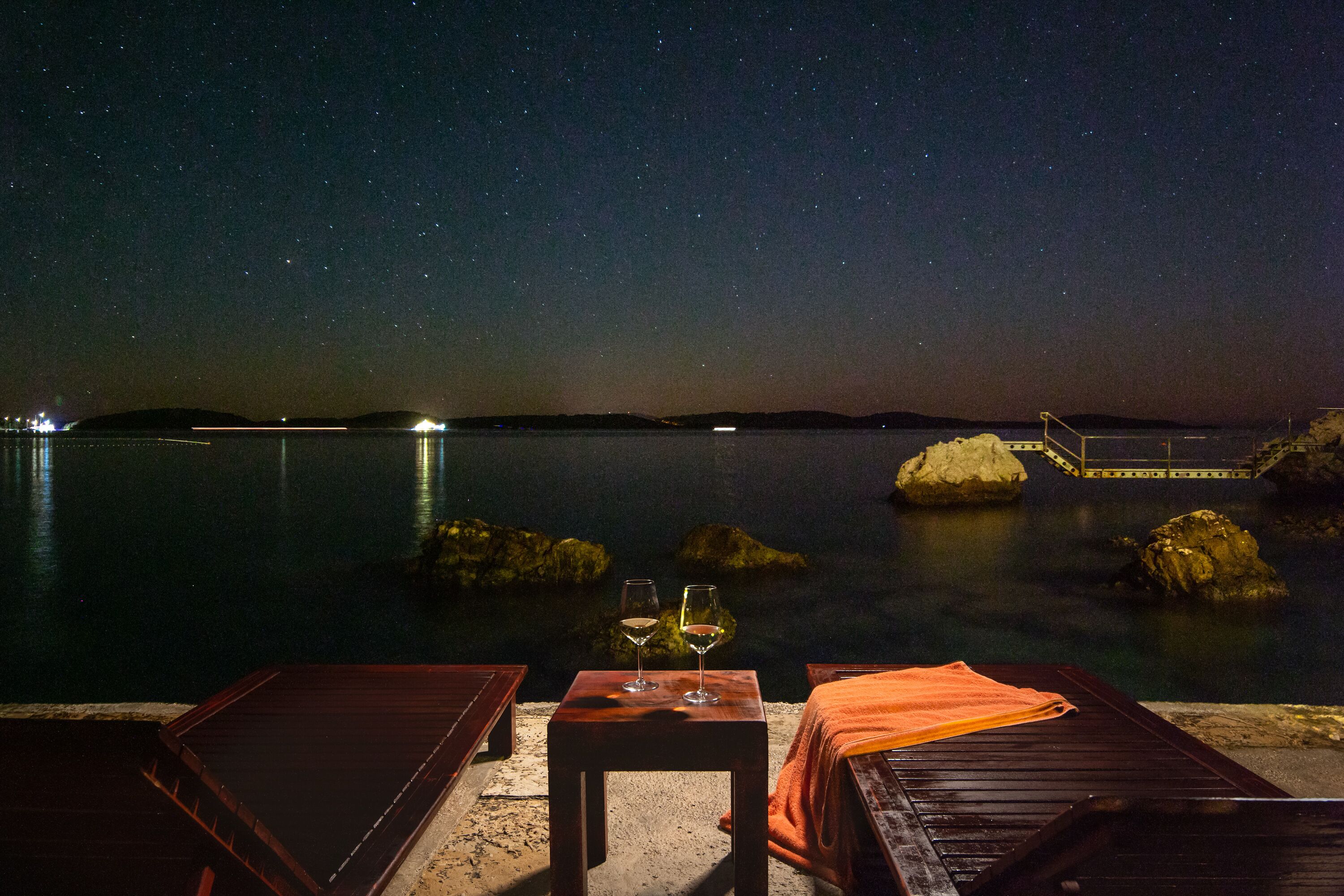 Une vue nocturne depuis une terrasse, avec deux verres de vin posés sur une table, donnant sur une étendue d'eau calme et des collines lointaines.