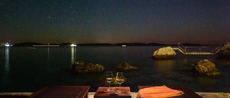 A night-time view from a terrace with two glasses of wine on a table, overlooking a calm body of water and distant hills.
